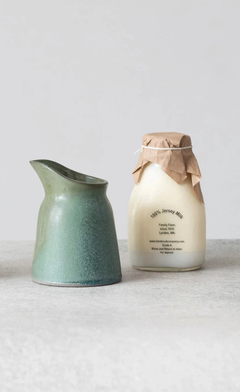 A matte green stoneware creamer with a spout, next to a milk bottle with a brown paper cover, on a neutral background.