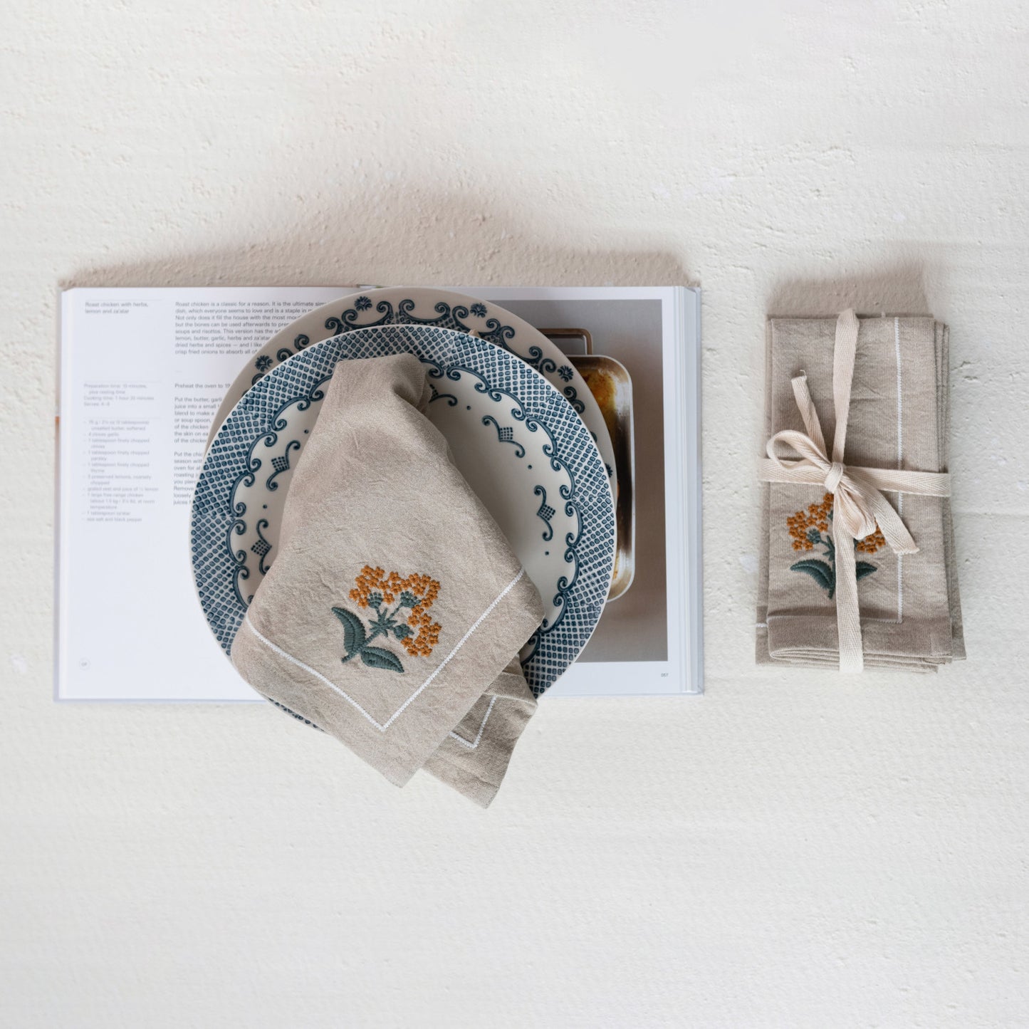 A set of beige cloth napkins with floral embroidery, displayed on a table alongside a plate and a book.