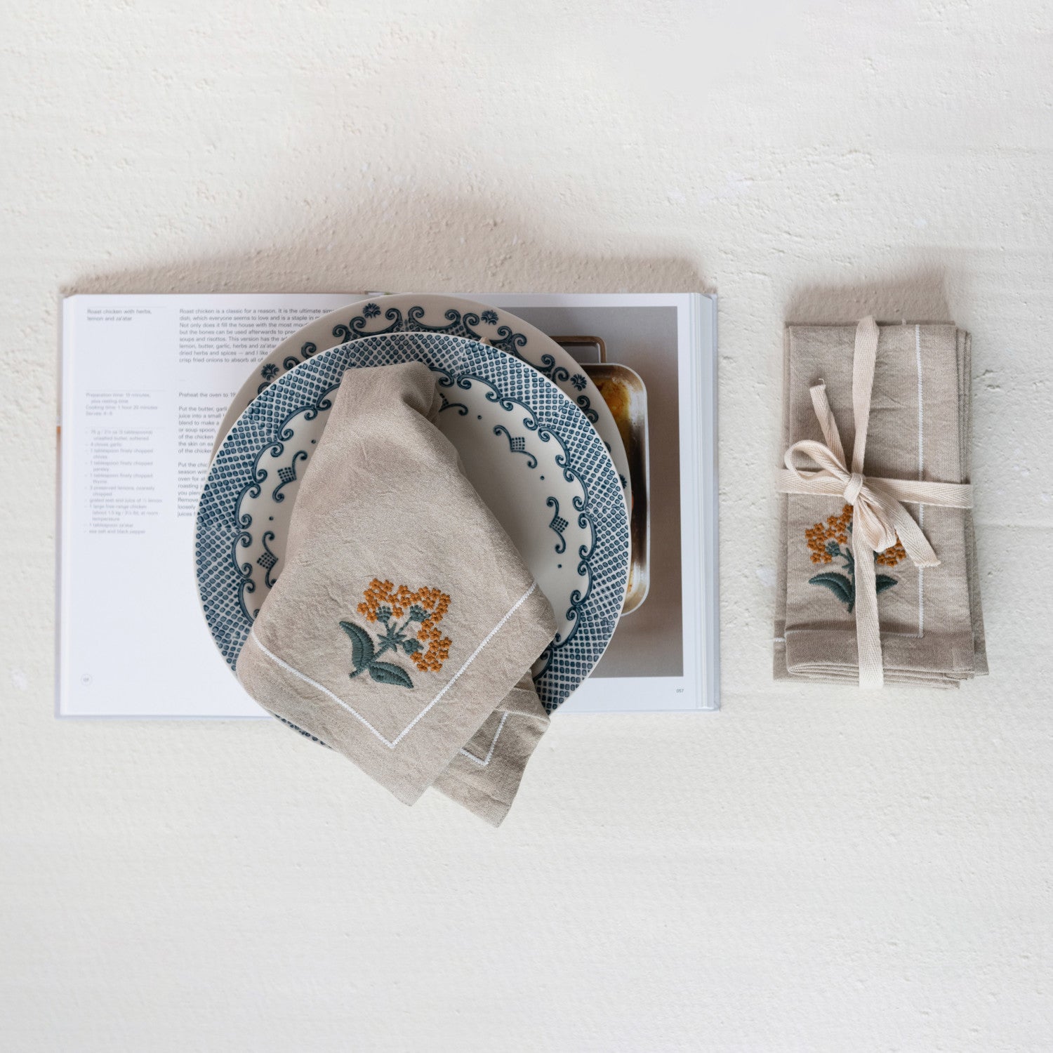 A set of beige cloth napkins with floral embroidery, displayed on a table alongside a plate and a book.