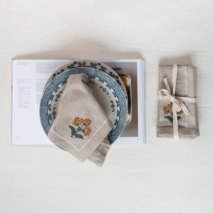 A set of beige cloth napkins with floral embroidery, displayed on a table alongside a plate and a book.