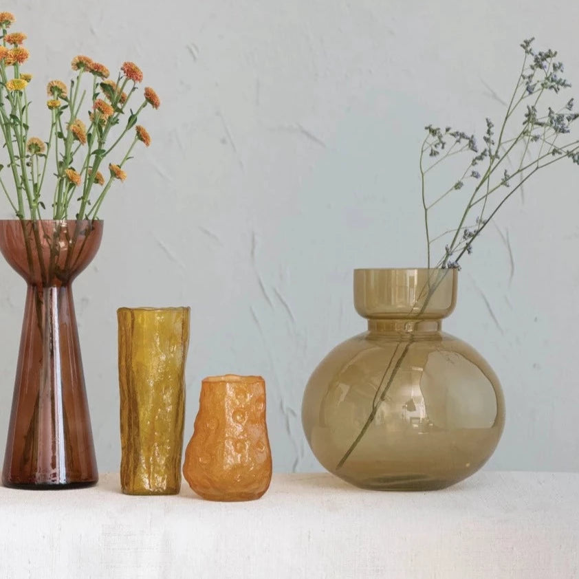 wheat glass vase with twiggy flowers in it arranged with other vase vases on a table.