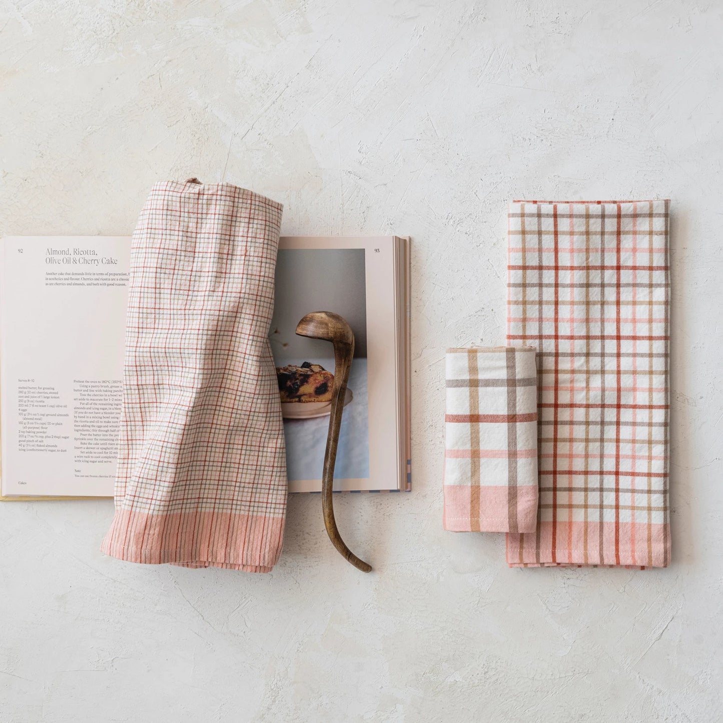 3 assorted plaid dishtowels folded and arranged on a table with an open cookbook and wooden ladle.