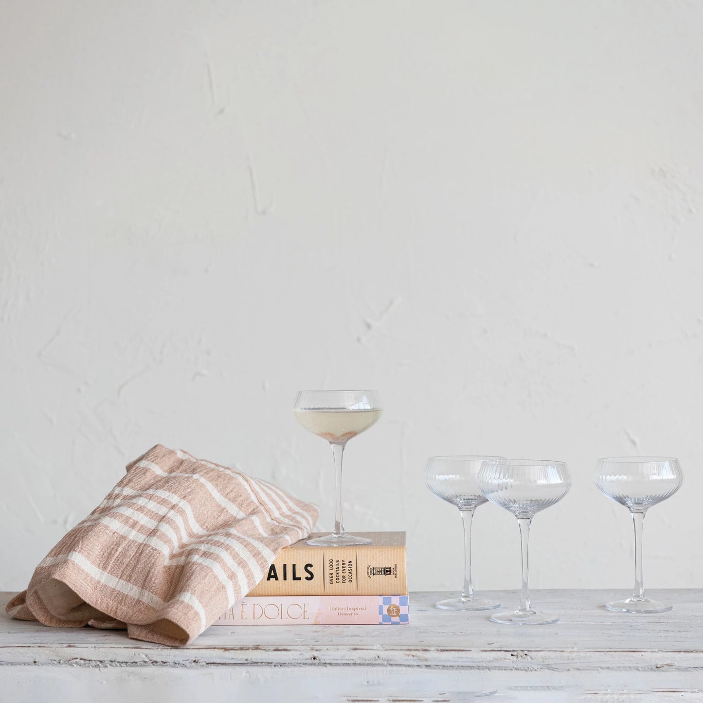 4 coupe glasses arranged on a wooden table with books and a tea towel.