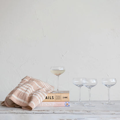 4 coupe glasses arranged on a wooden table with books and a tea towel.