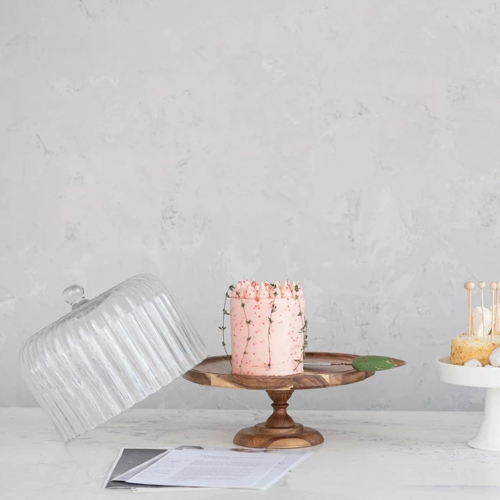 wooden pedestal with pink cake on it and fluted glass dome set askew on the pedestal.
