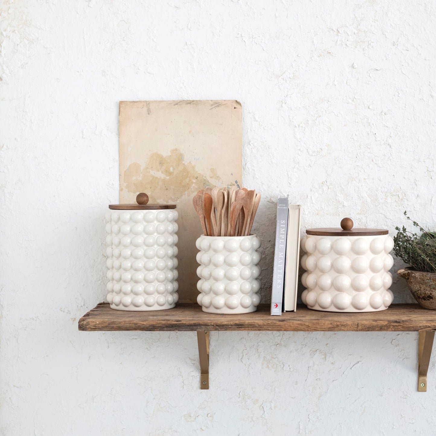 all three sizes of raised dots and acacia wood lid stoneware canisters displayed on a wooden shelf hanging on a white wall with two books and bowls with greenery