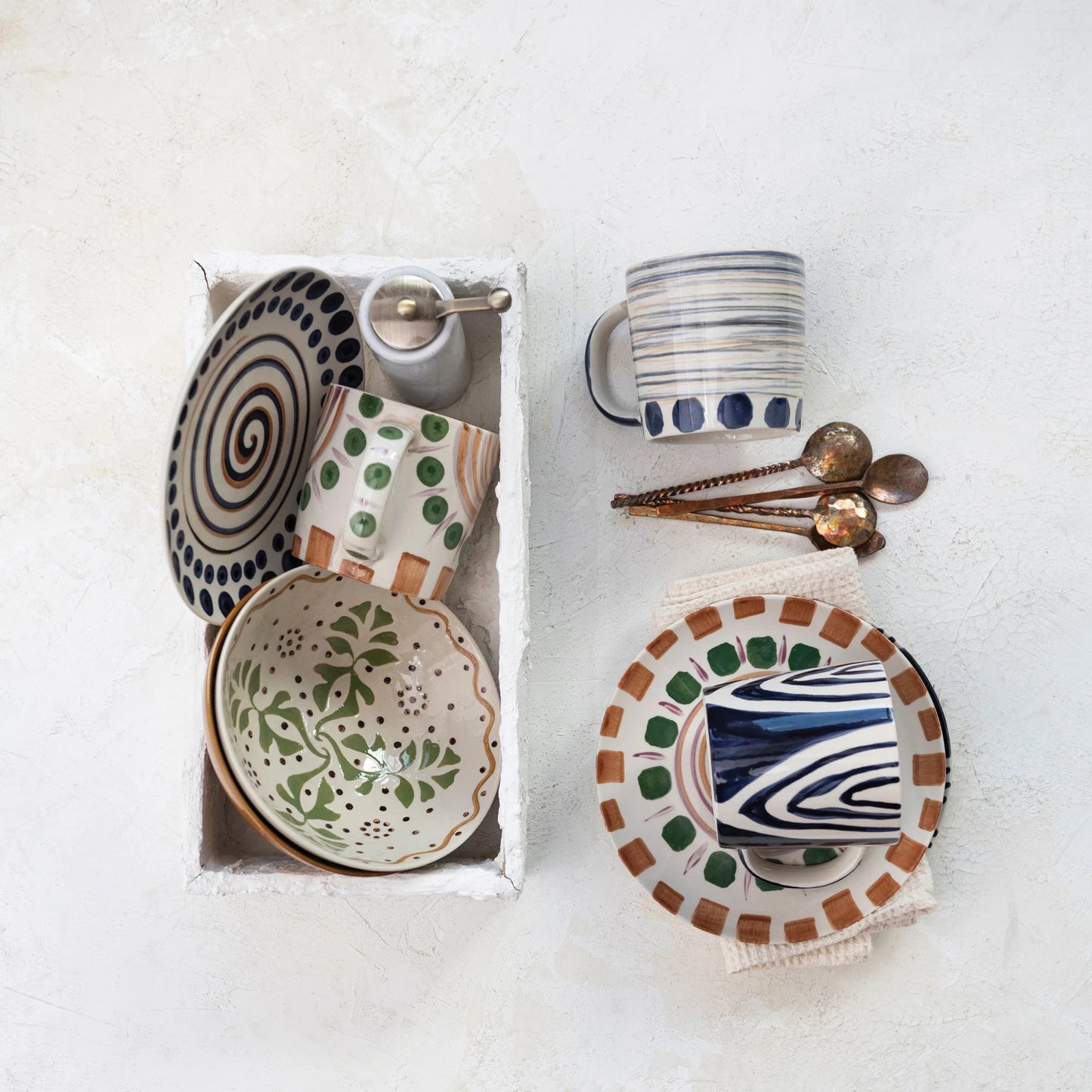 top view of all three styles of hand painted stoneware mugs displayed with plates, salt grinder, hammered spoons, and a white box on a white surface