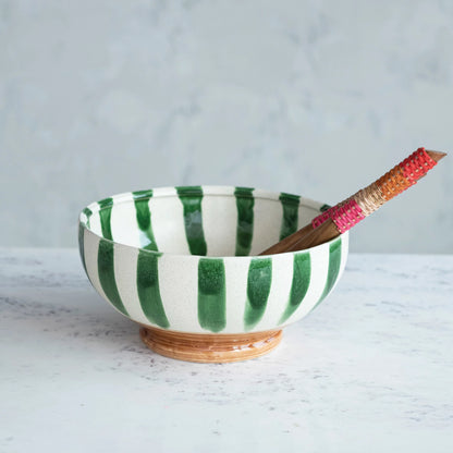 cream stoneware bowl with green vertical stirpes and brown base set on a grey table with colorful serving utensils in it.