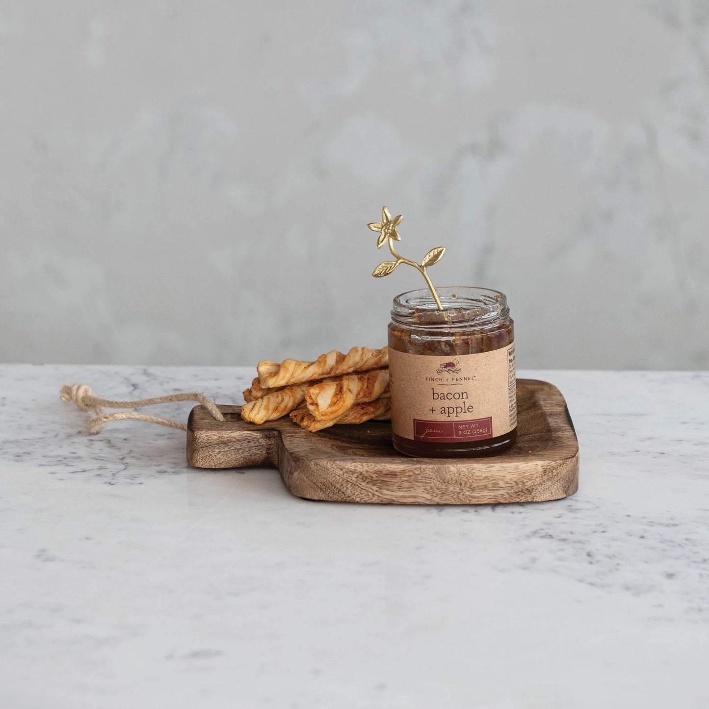 mango wood board on a marble counter with a jar of jam and bread sticks on it.