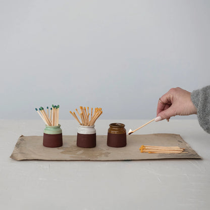 all three colors of stoneware match holder with striker filled with matches and displayed on a torn recycled paper with a womans hand holding a lit match