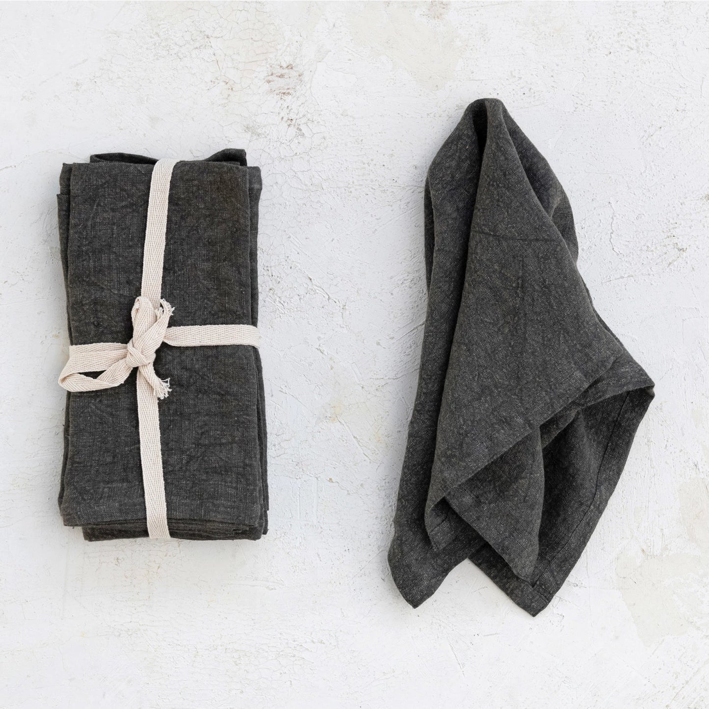 set of charcoal napkins tied together with a ribbon and one napkin draped next to it on a plaster background.