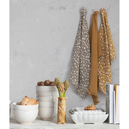 both colors of floral printed cotton waffle tea towels hanging on a wall behind stacked bowls, vase with flowers, and books