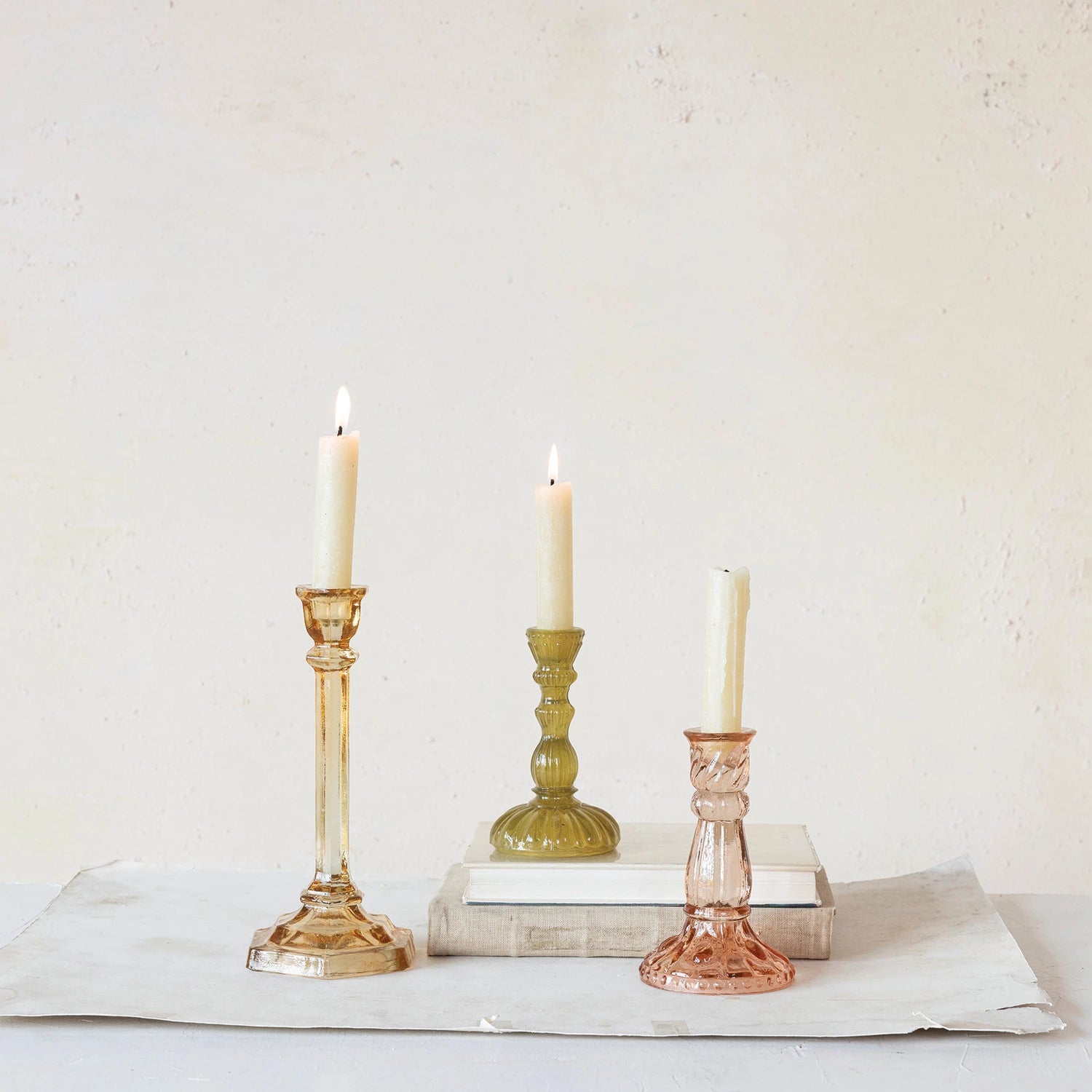 3 styles of glass candle holders with off-white tapers in them arranged on a counter with books.