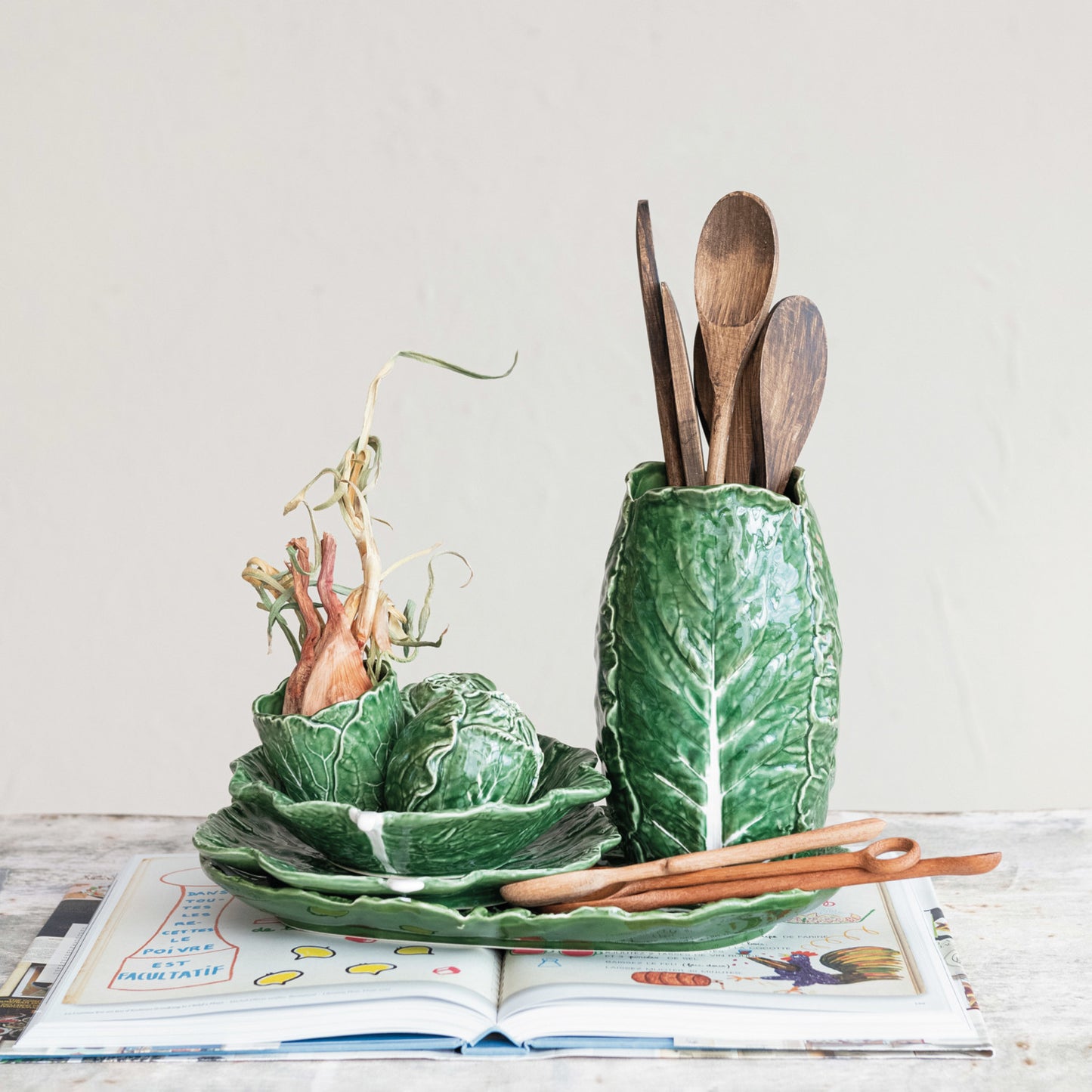 cabbage leaf vase filled with wooden utensils set on a table with other cabbage leaf shaped dishes.