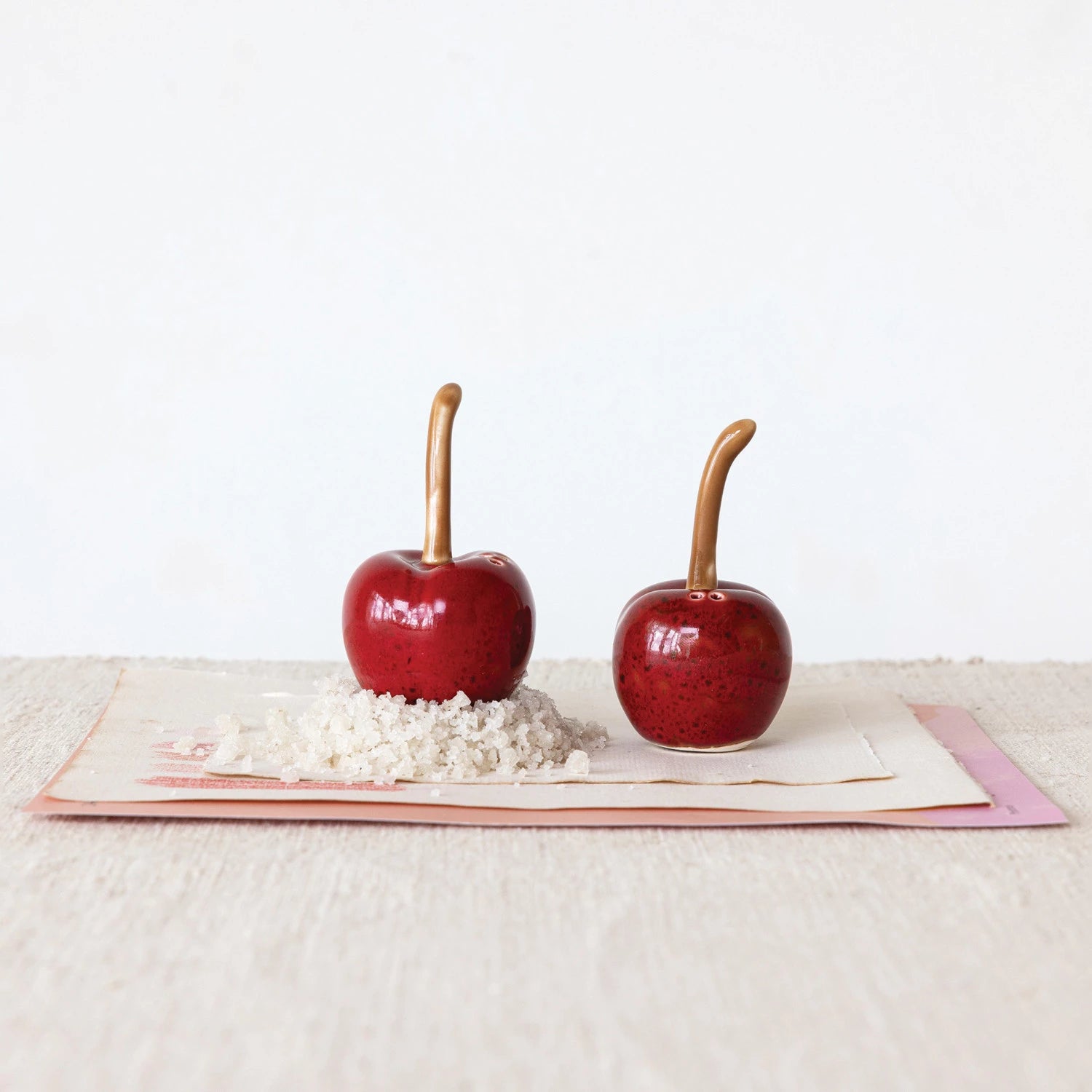 2 cherry shaped shakers on a table, one shaker is set on a pile of salt.