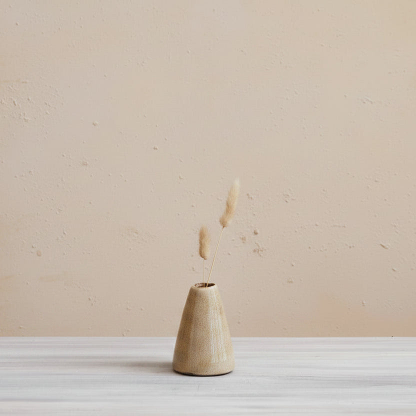 small tan bud vase with 2 stems of dried grasses in it set in front of a plaster wall