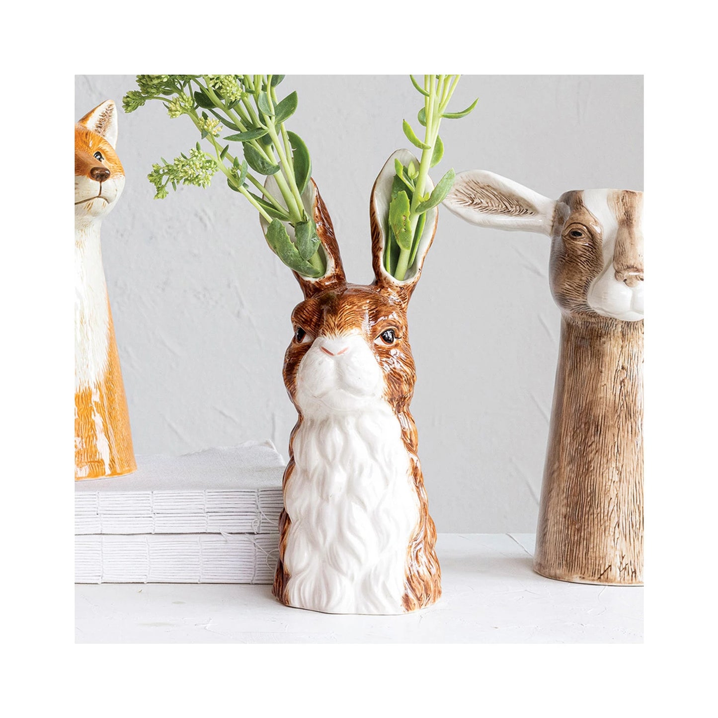 Vase shaped like a rabbit with greenery in it surrounded by other vases, on a white surface