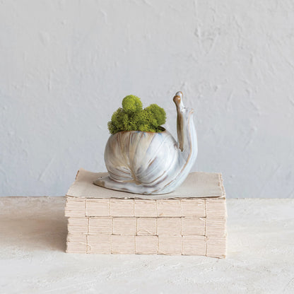 A stoneware snail-shaped planter with a green plant on its back, placed on a stack of books against a neutral background.