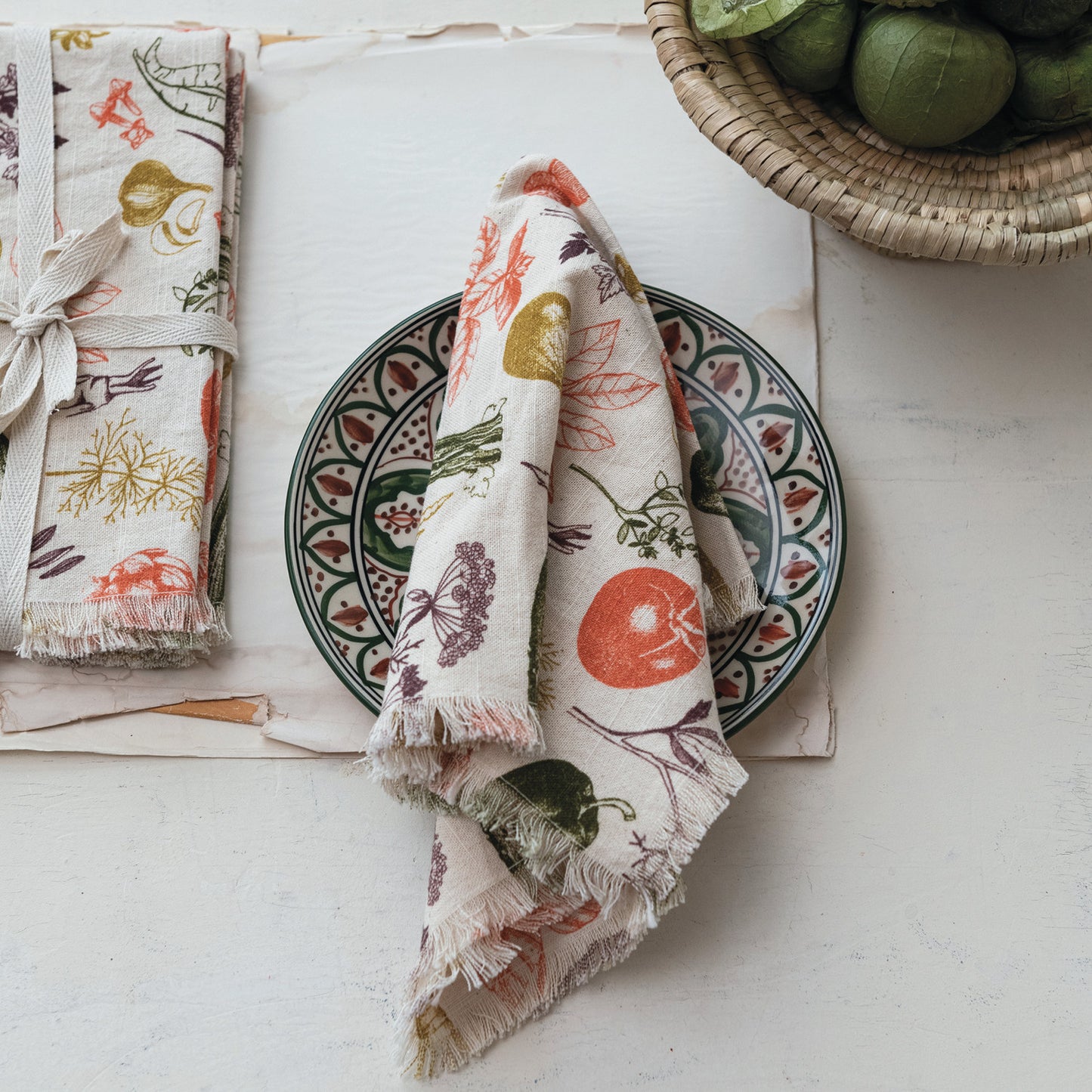 Veggie Printed Napkin draped over a plate set on a table with other napkins and a bowl of fruit.