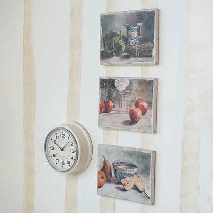 three framed still life paintings displayed on a wall alongside a round white clock.
