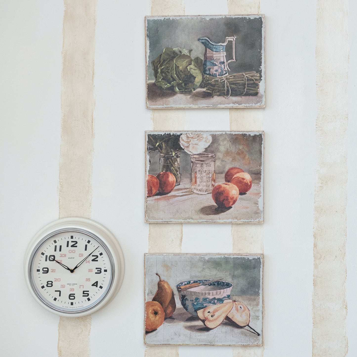 3 still-life artworks hanging in a vertical row on a stiped wall with a  round white clock