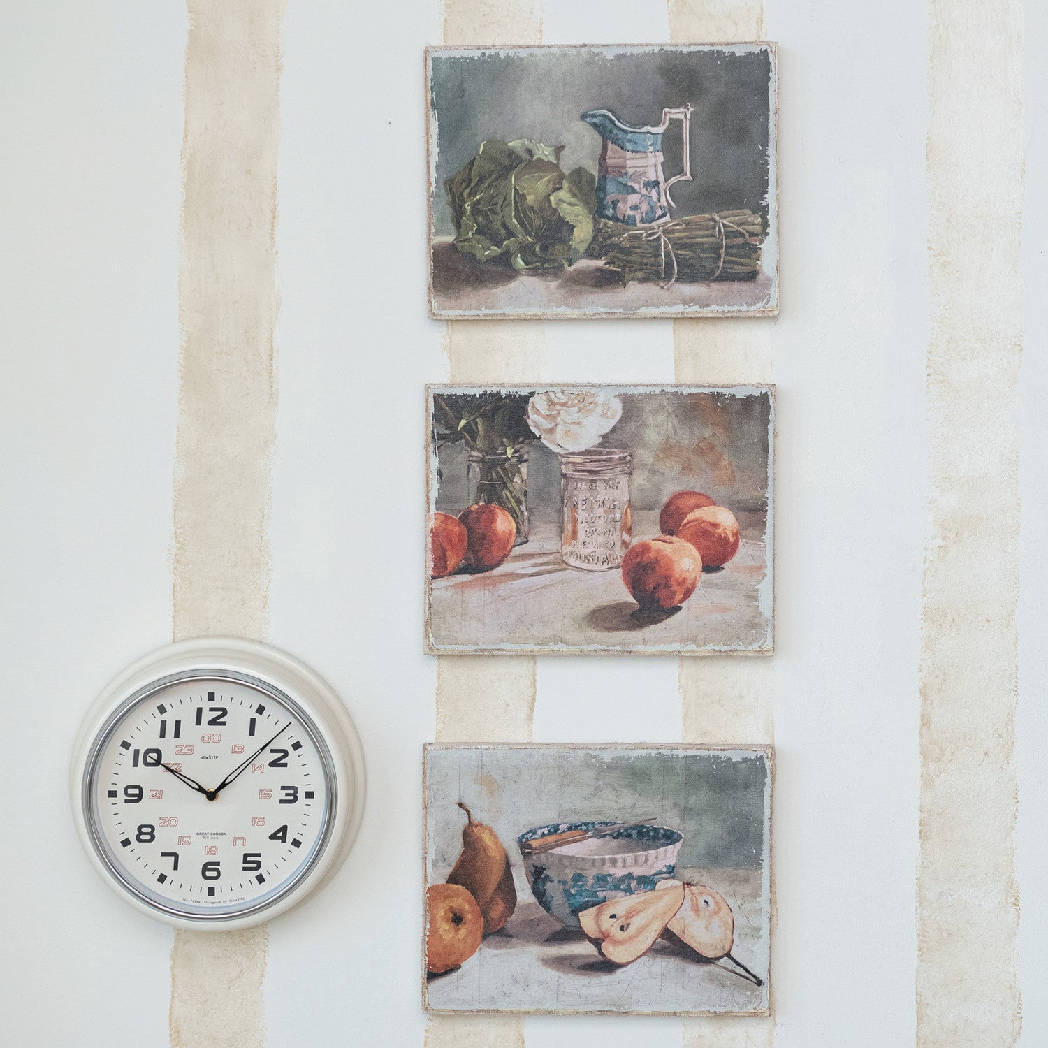 3 still-life artworks hanging in a vertical row on a stiped wall with a  round white clock