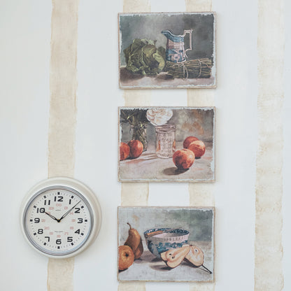 3 still-life artworks hanging in a vertical row on a stiped wall with a  round white clock