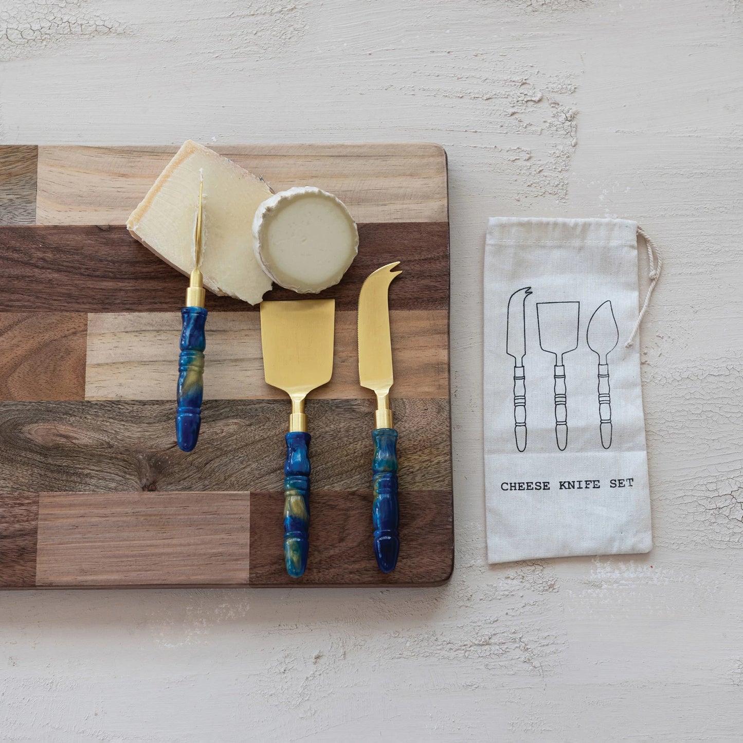 Cheese knife set with blue handles on a wooden cutting board with cheese, arranged on a plaster surface