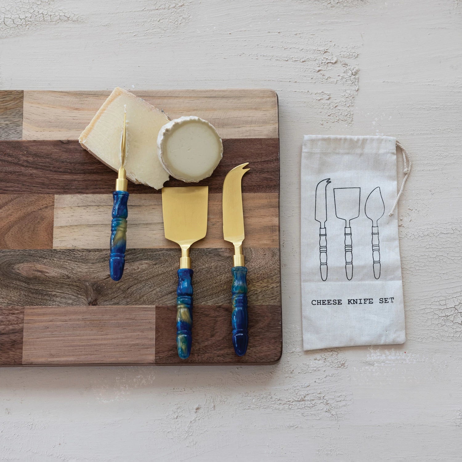 Cheese knife set with blue handles on a wooden cutting board with cheese, arranged on a plaster surface