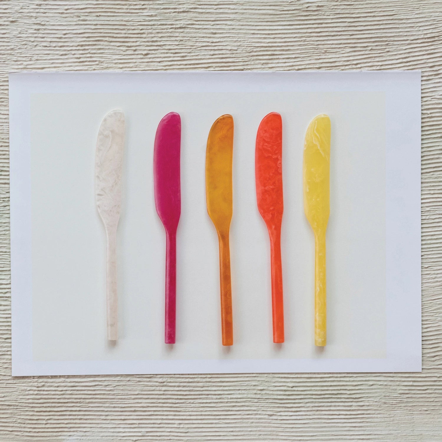 Five colorful butter knives on a white card with a wooden surface background