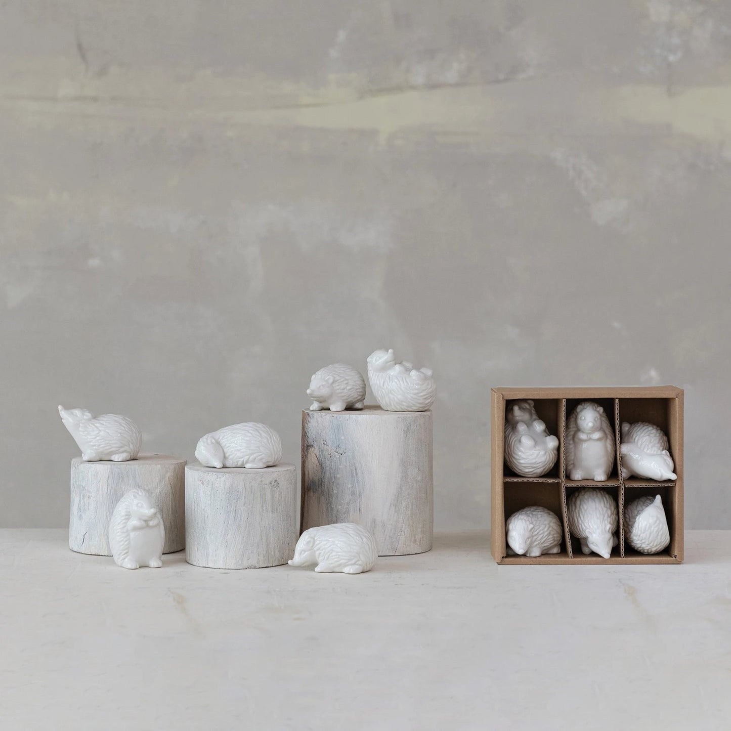 Decorative ceramic hedgehogs on cylindrical bases with a wooden box containing more pieces against a gray background.