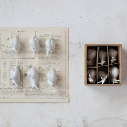 6 assorted white ceramic snails arranged on a printed piece of paper with a boxed set of snails set next to it