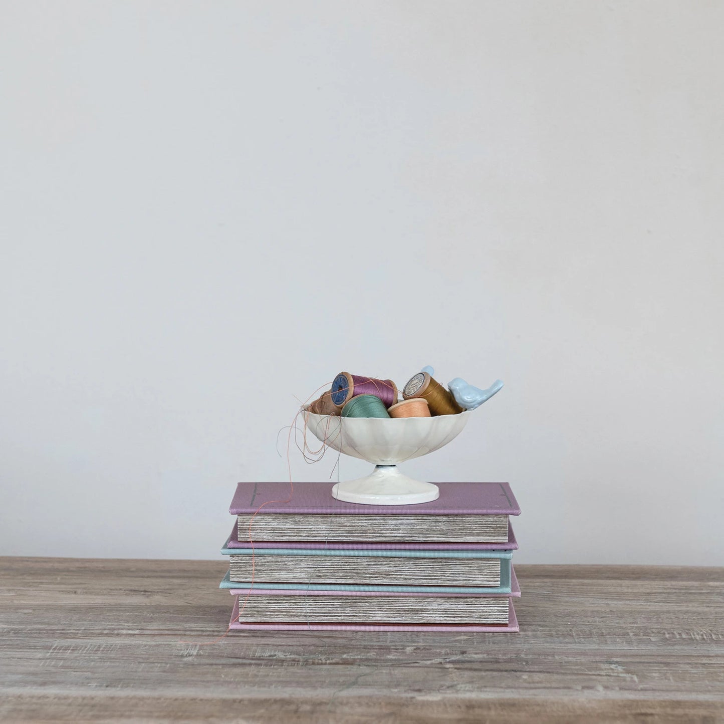 Stack of books with a decorative bowl containing colorful spools of thread on a wooden surface.