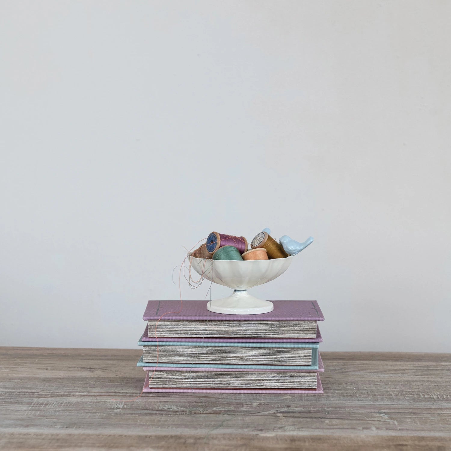Stack of books with a decorative bowl containing colorful spools of thread on a wooden surface.