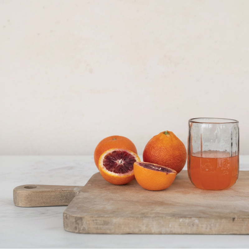 short drinking glass partially filled with juice set on a board with 3 oranges