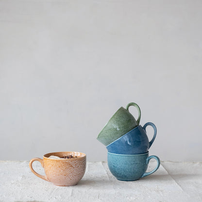 Three ceramic mugs in different colors stacked on a light surface and a coral colored mug set next to them on a grey background