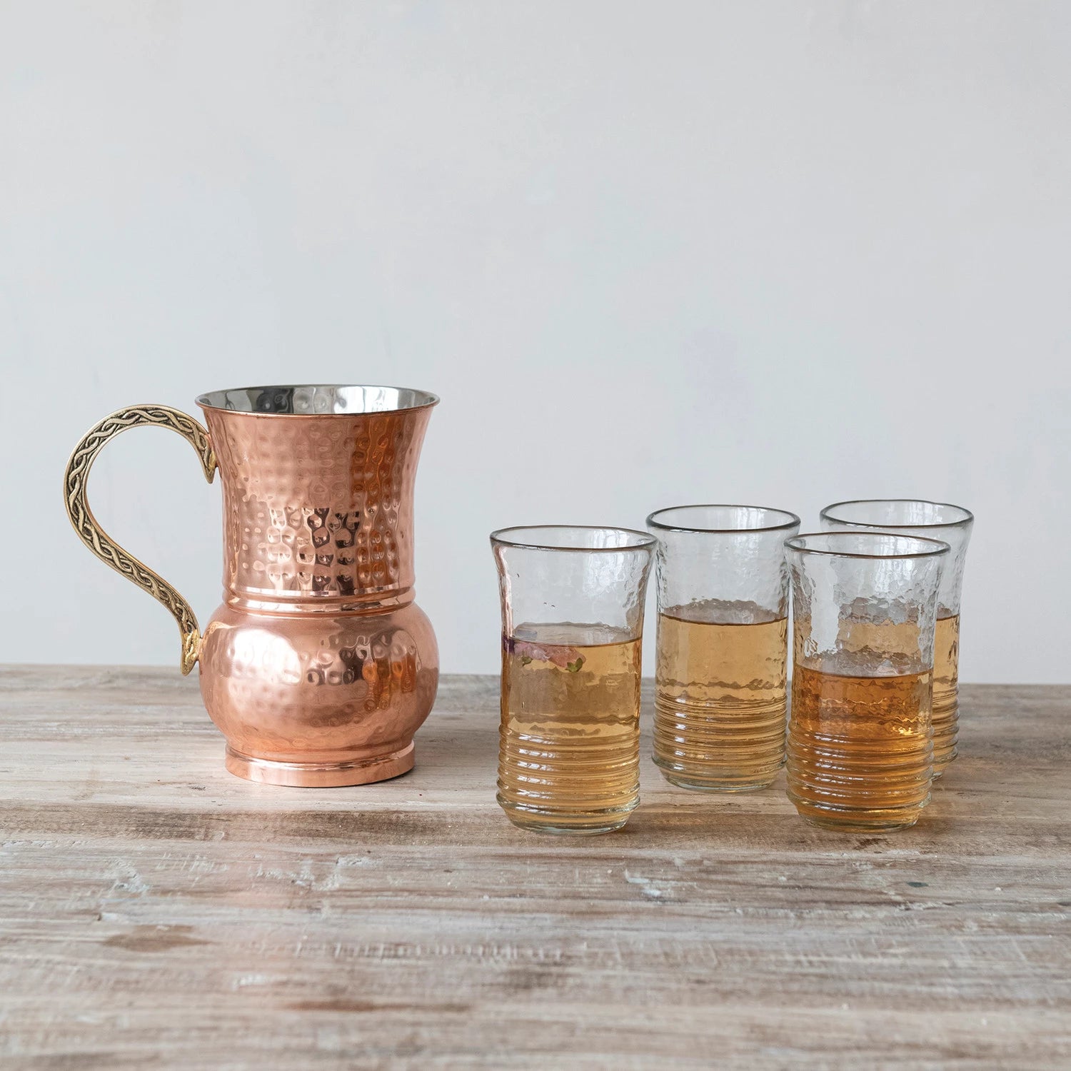 Copper pitcher and drinking glasses filled with beverage set on a wooden surface with a light background