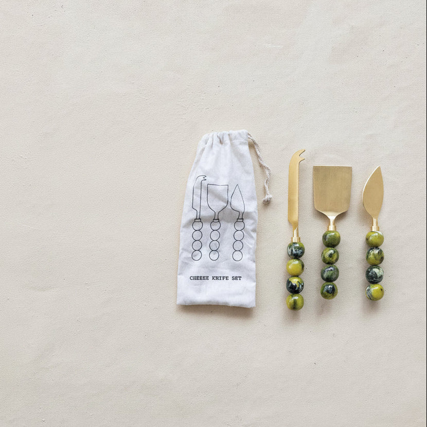 Set of gold cheese serving tools with green beaded handles on a beige background