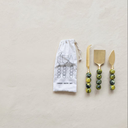 Set of gold cheese serving tools with green beaded handles on a beige background