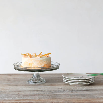 cake on a glass stand with plates and a fork on a wooden surface.