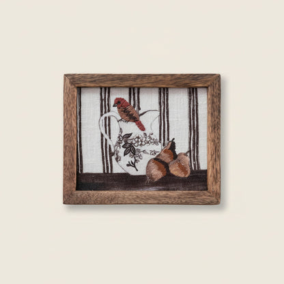 Framed art with embroidered birds sitting on a floral pitcher with pears next to it and brown striped background