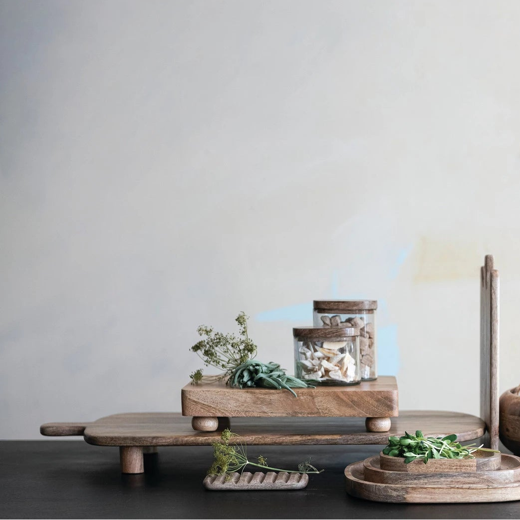 wooden kitchen items including a cutting board, bowls, and jars scattered with fresh herbs on a dark surface with a light gray background.