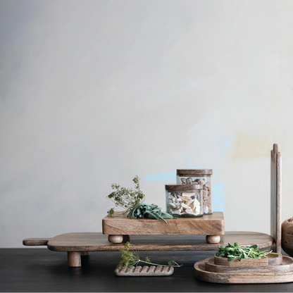 wooden kitchen items including a cutting board, bowls, and jars scattered with fresh herbs on a dark surface with a light gray background.