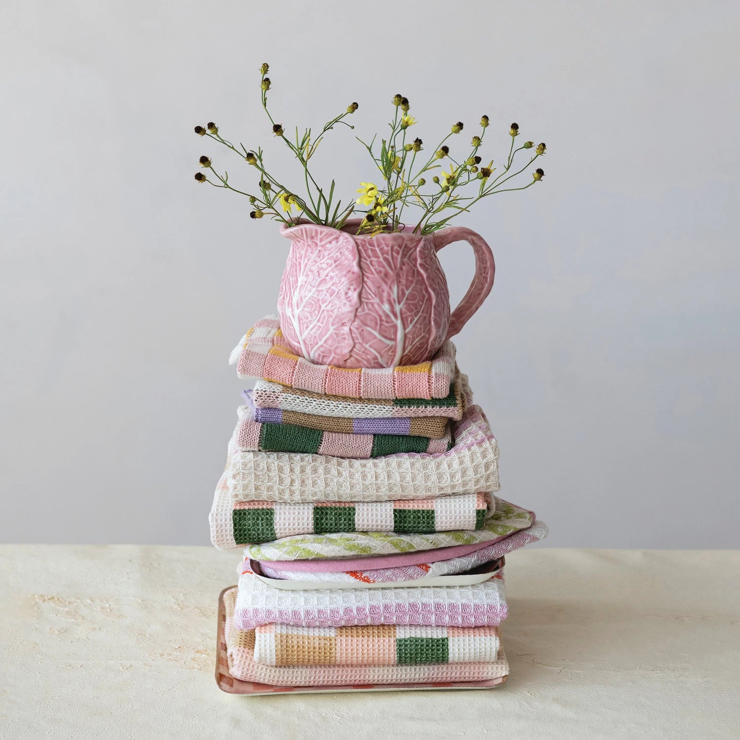 Stack of colorful towels with a pink cabbage cream pitcher holding flowers on a light background