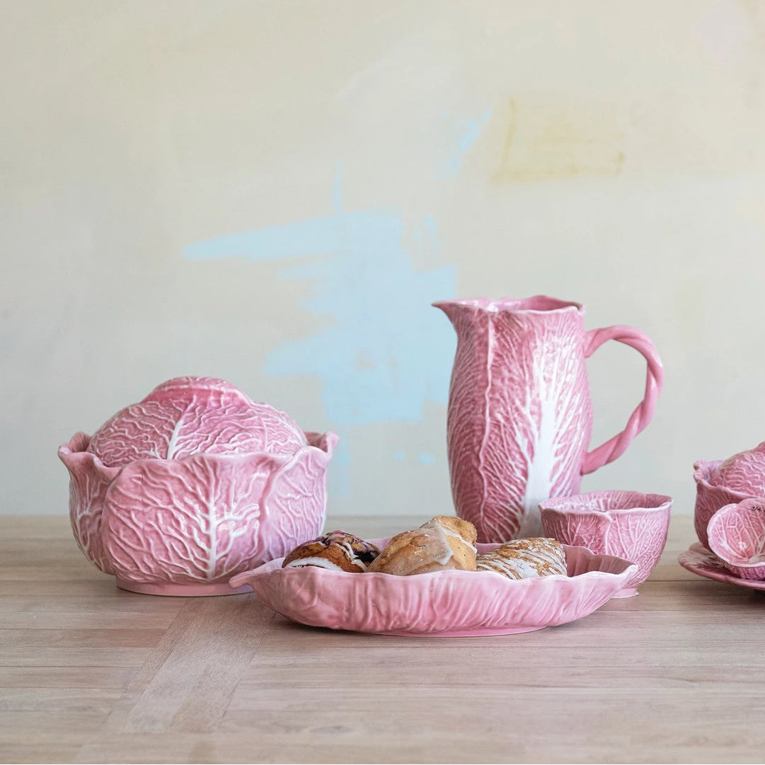 pink ceramic kitchenware including a tureen, cups, and a pitcher all shaped like cabbage arranged on a wooden surface.