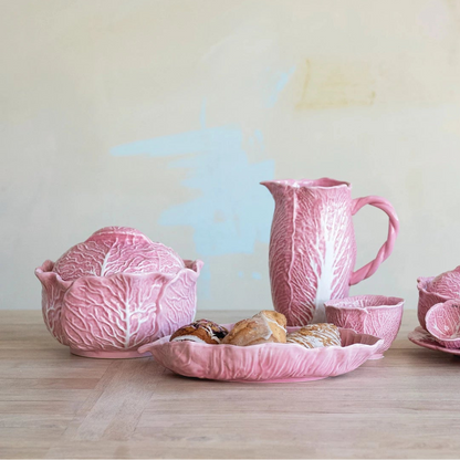 pink ceramic kitchenware including a tureen, cups, and a pitcher all shaped like cabbage arranged on a wooden surface.