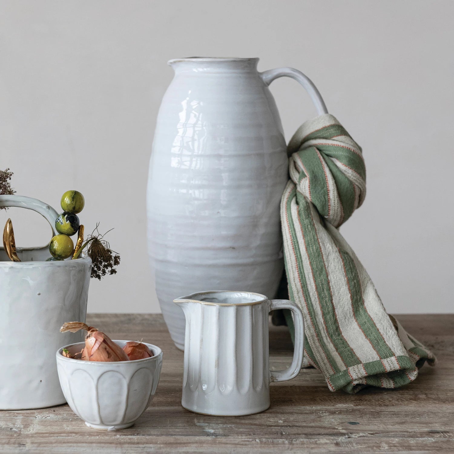 Ceramic pitcher, mug, and small bowl on a wooden surface with a striped towel draped over the pitcher.