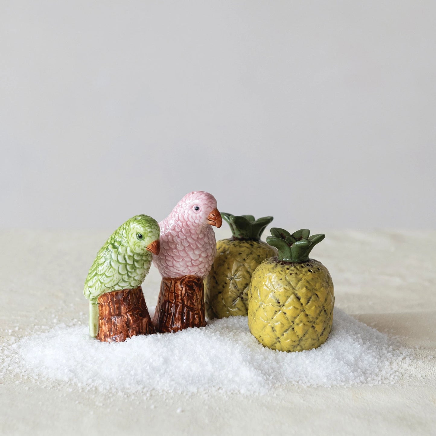 Decorative salt and pepper shakers shaped like birds arranged with ceramic pineapples, all set in a pile of loose salt