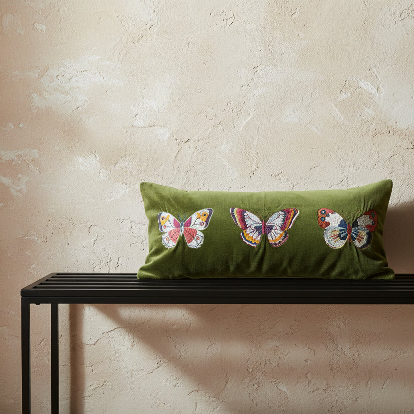 An olive green rectangular pillow with three colorful embroidered butterflies, resting on a black metal bench against a textured beige plaster wall.
