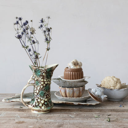 embossed Ceramic pitcher with grape and leaf pattern with dried stems in it set on a wooden table with a stack of dishes